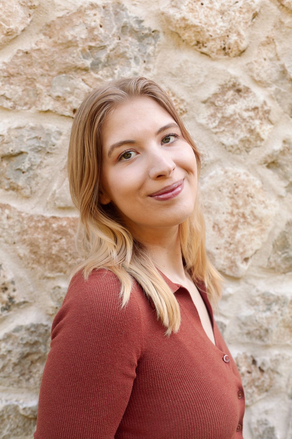 Blonde woman in a rust ribbed top smiling against a stone wall background.