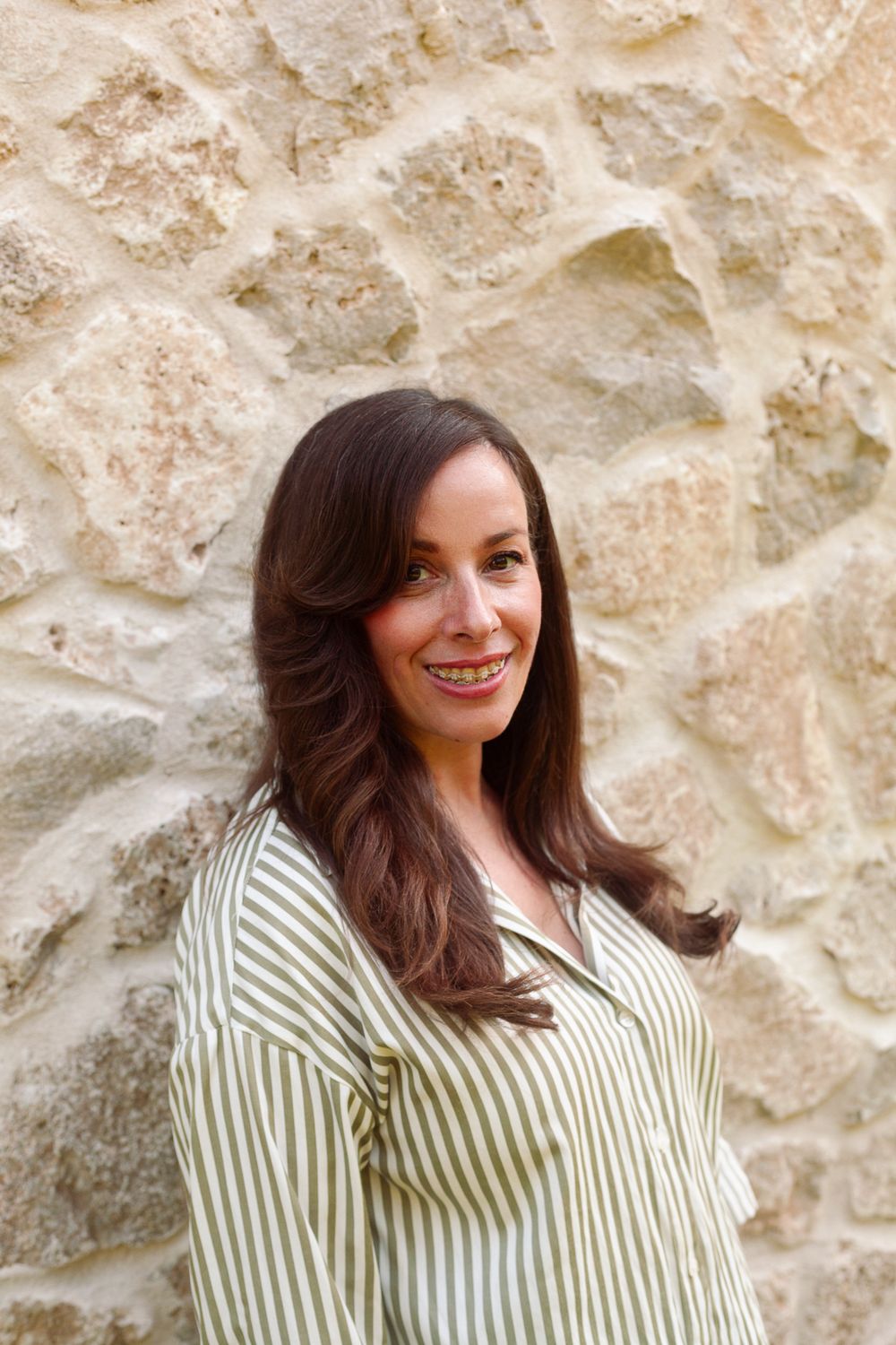 Brunette woman in a green striped shirt smiling warmly against a stone wall.