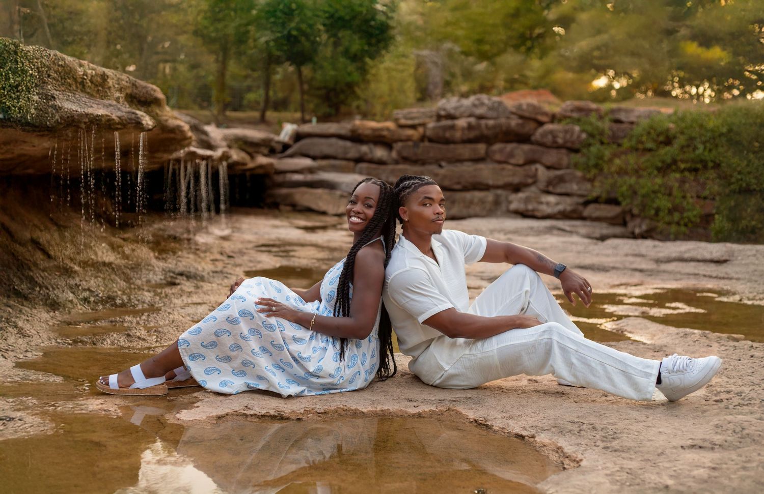 A couple in white outfits sits together on red rocks near a stream during a romantic outdoor photoshoot.
