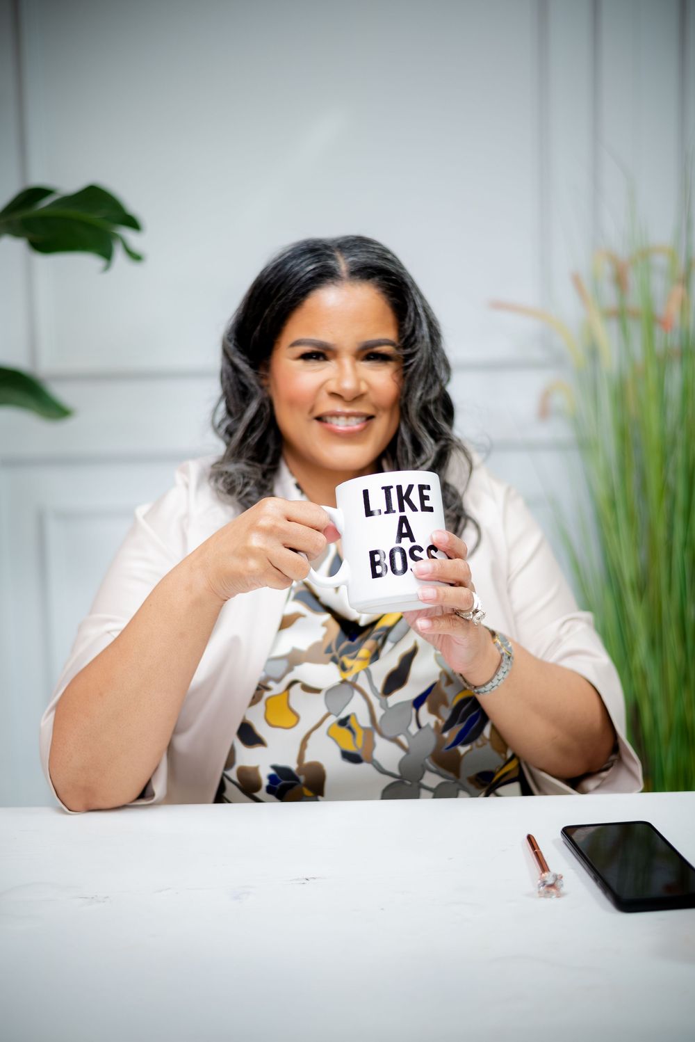 Professional wearing white blouse holds up paper signs with text while smiling at desk with office plants in background.