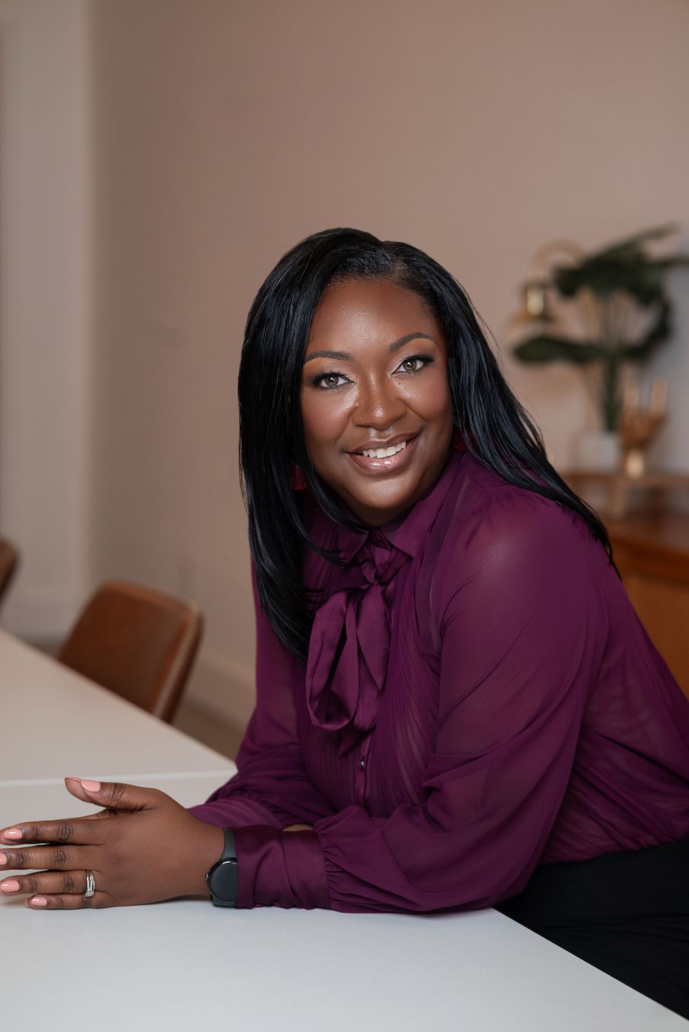 Professional portrait of someone in a burgundy blouse with a bow tie collar smiling warmly while seated at a white table.