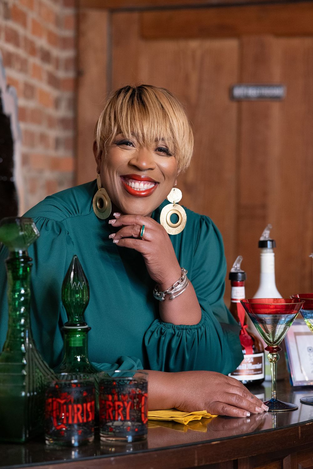 Person in teal top smiles while sitting at a bar counter with various bottles and drinks displayed.