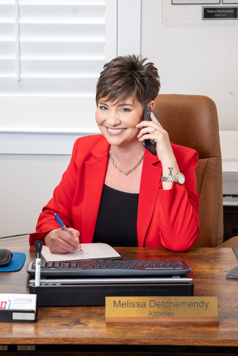 Professional in red blazer works at wooden desk in office setting with nameplate and computer.
