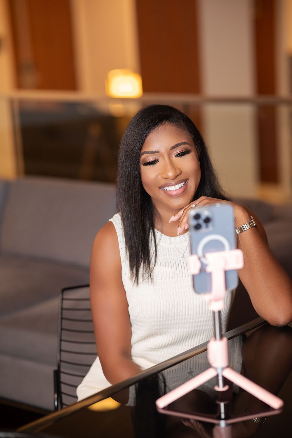 Content creator recording with a smartphone on a tripod setup while wearing a white sleeveless top in warm indoor lighting.