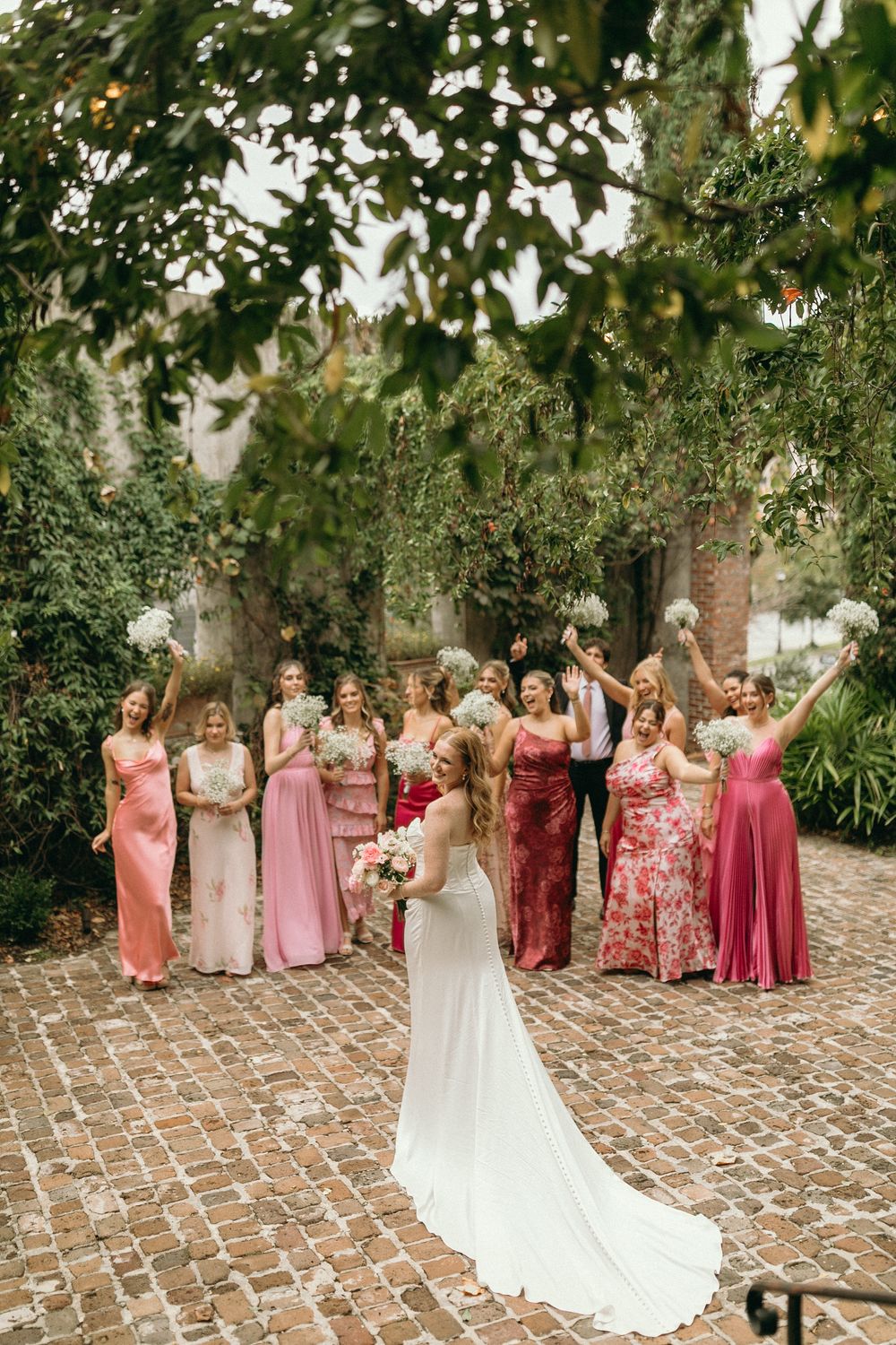 Wedding party wearing pink and floral bridesmaid dresses pose together on brick patio with greenery backdrop at Summerour Studio