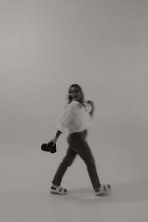 A blurred motion shot of a photographer in white shirt and jeans walking with camera gear against neutral background.