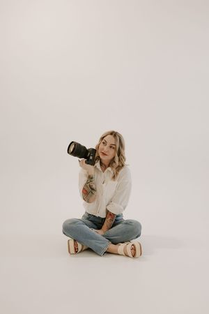 Photographer in white shirt and jeans poses playfully with camera against white background.
