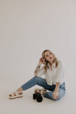 Model in white shirt and jeans poses casually on floor against neutral backdrop.