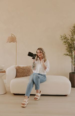 A photographer sits on white couch holding camera, wearing casual white shirt and jeans in minimal studio setting.