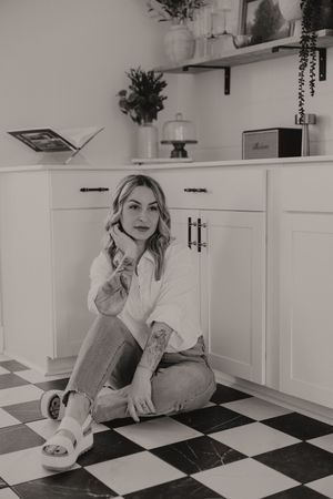 Black and white photo of photographer sitting on checkered floor against white kitchen cabinets.