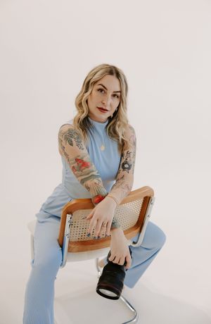Model in light blue dress poses with camera in vintage cane chair against white backdrop.