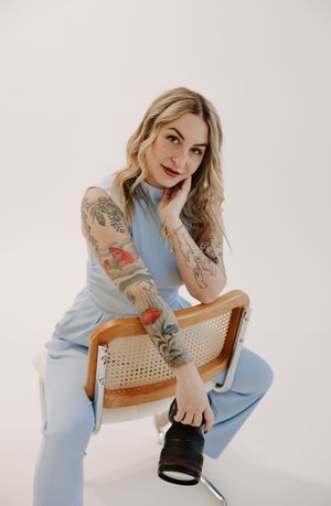 A person in a light blue outfit poses on a wooden chair against a white backdrop showing tattoo art on their arms.