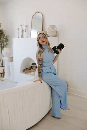 Professional photographer in blue dress poses with camera in minimalist white studio setting.