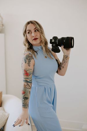 Photographer with tattoos holds camera while wearing light blue dress in bright studio.