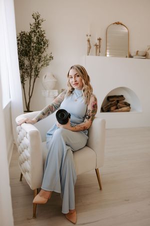 Photographer in blue dress demonstrates camera poses in bright minimalist studio setting.