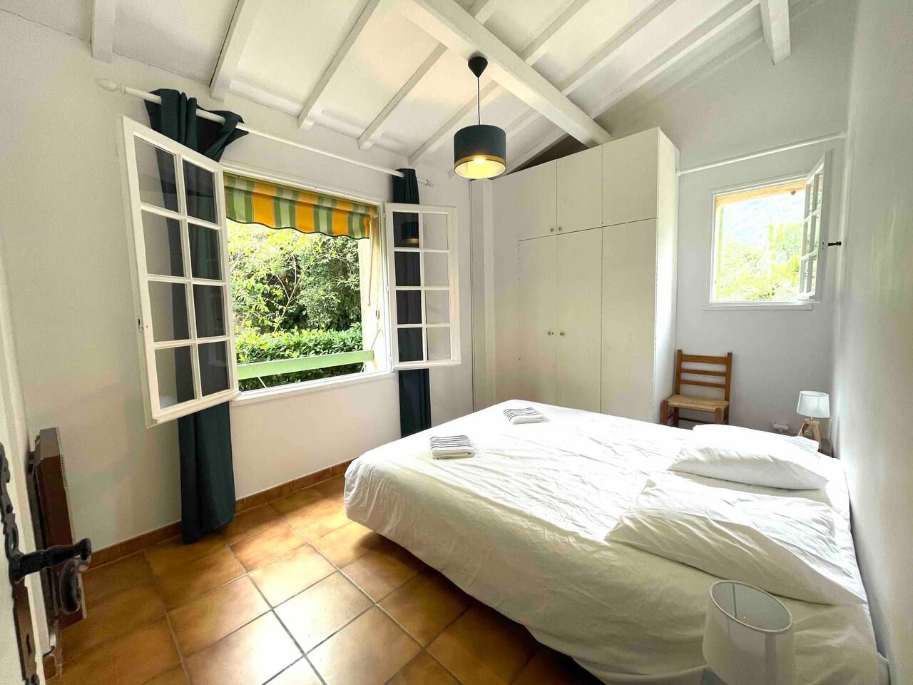 Bright and airy bedroom interior with white walls, wooden floors, and natural light streaming through multiple windows.