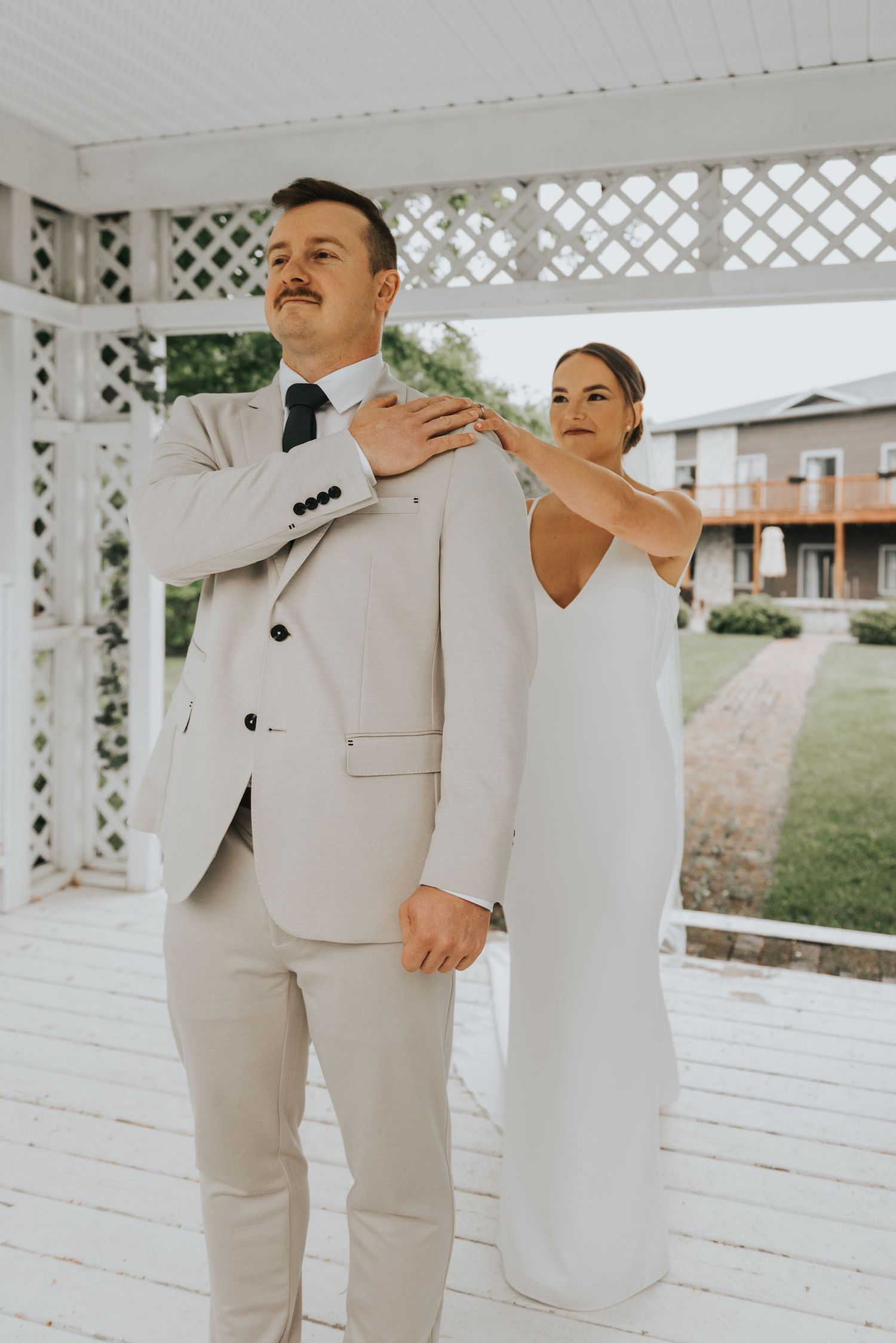 A couple in wedding attire playfully poses together under a white latticed gazebo during their outdoor ceremony.