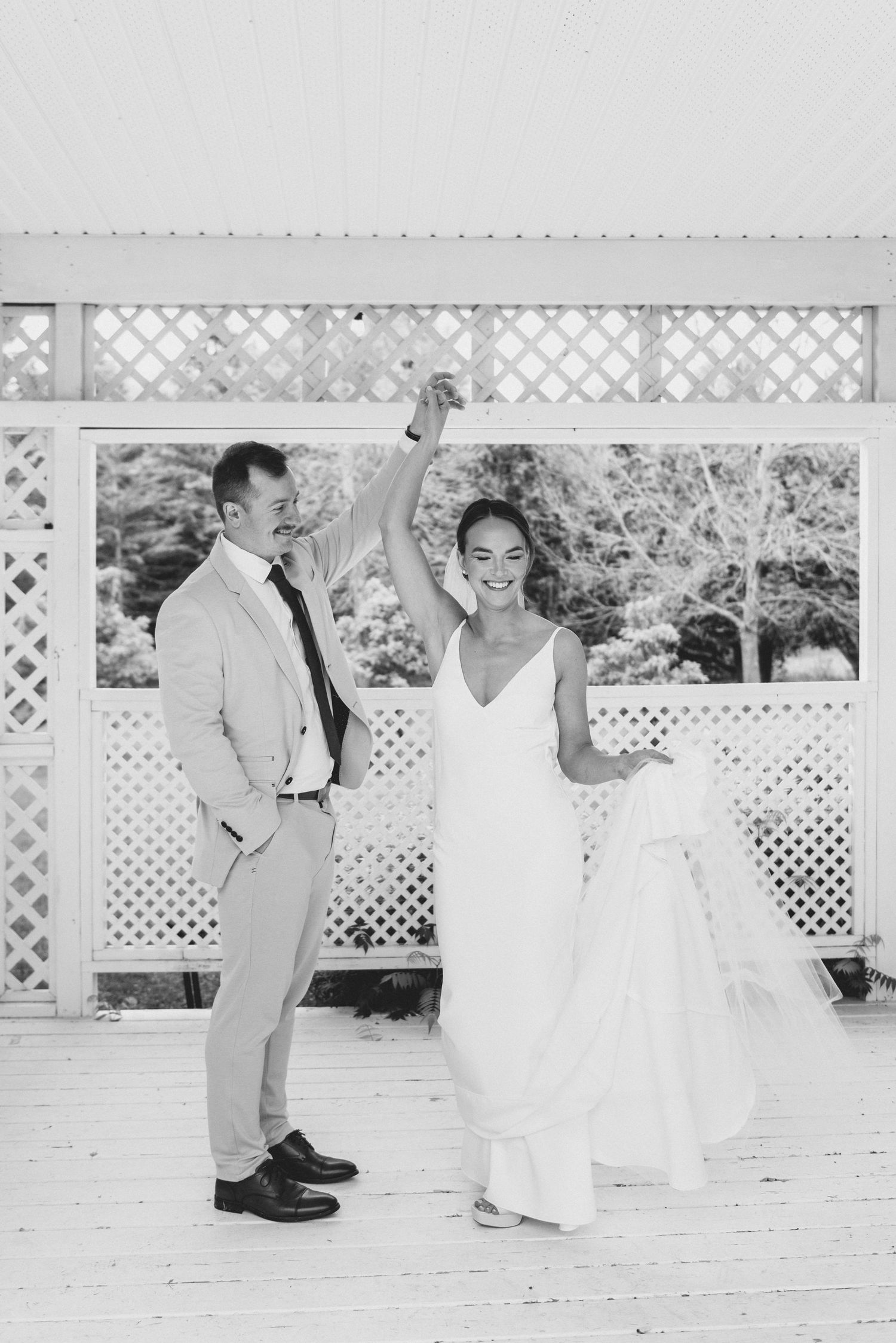 A couple in wedding attire dance together on a white lattice gazebo during their outdoor celebration.