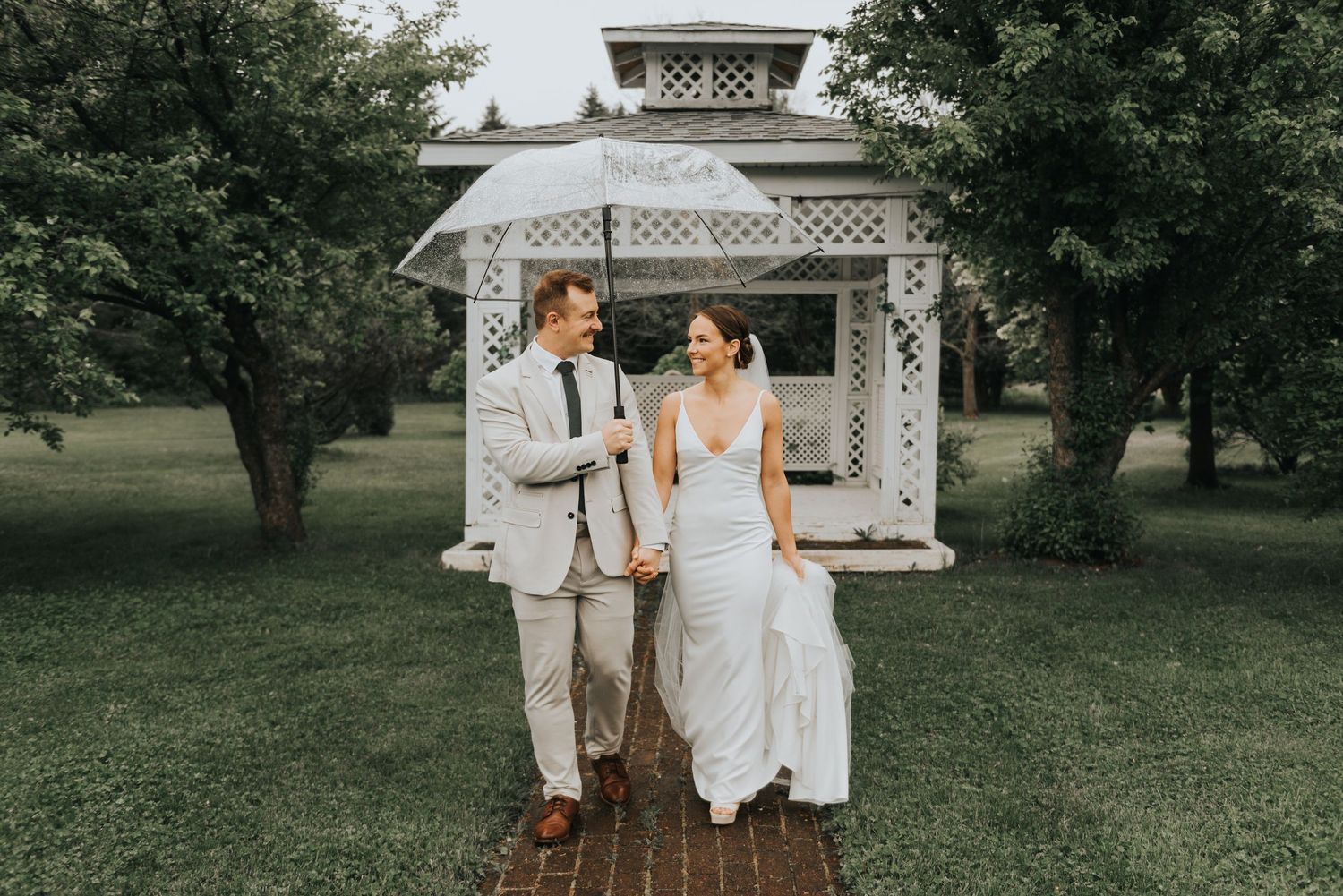 A couple shares a romantic moment in front of a white gazebo in a lush garden setting.