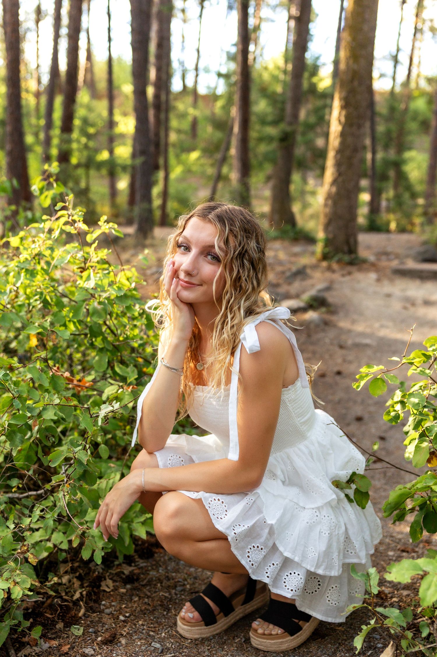 A person in a white eyelet dress sits thoughtfully among pine trees and greenery in a forest setting.