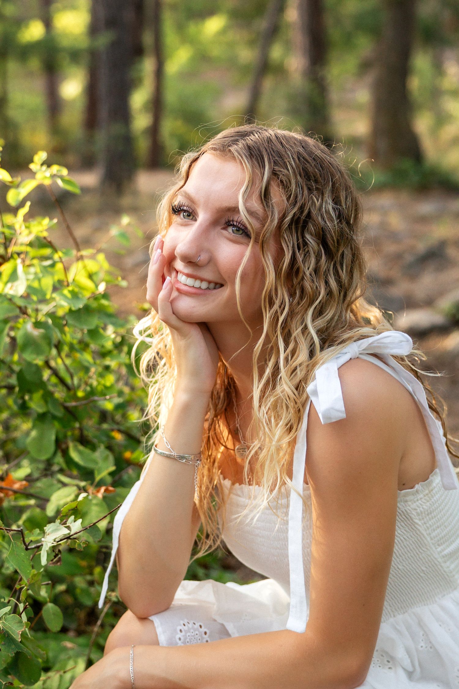 A woman in a white eyelet dress poses outdoors in dappled sunlight among green foliage.