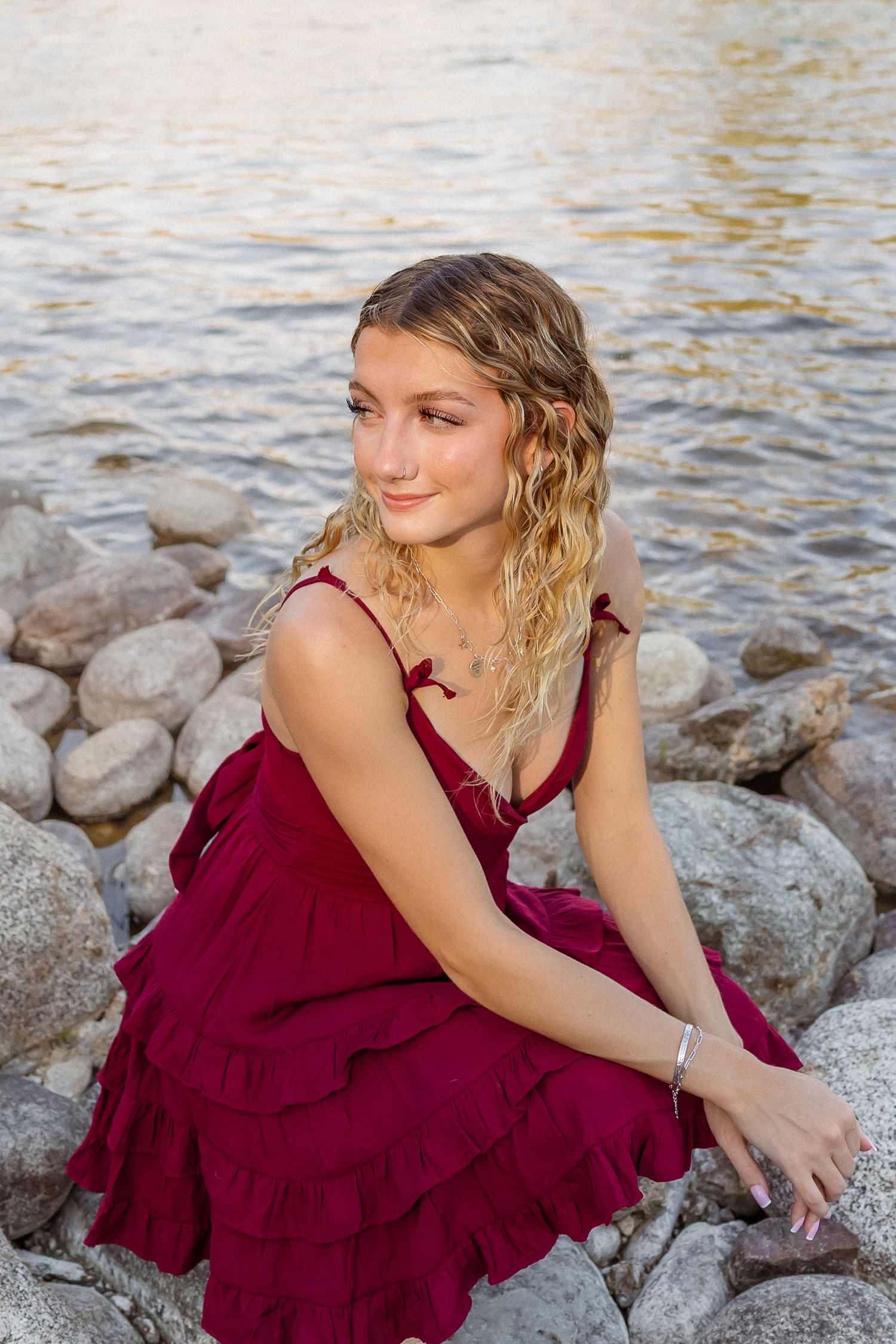 A person in a burgundy ruffled dress poses gracefully on rocky waterfront terrain during golden hour.