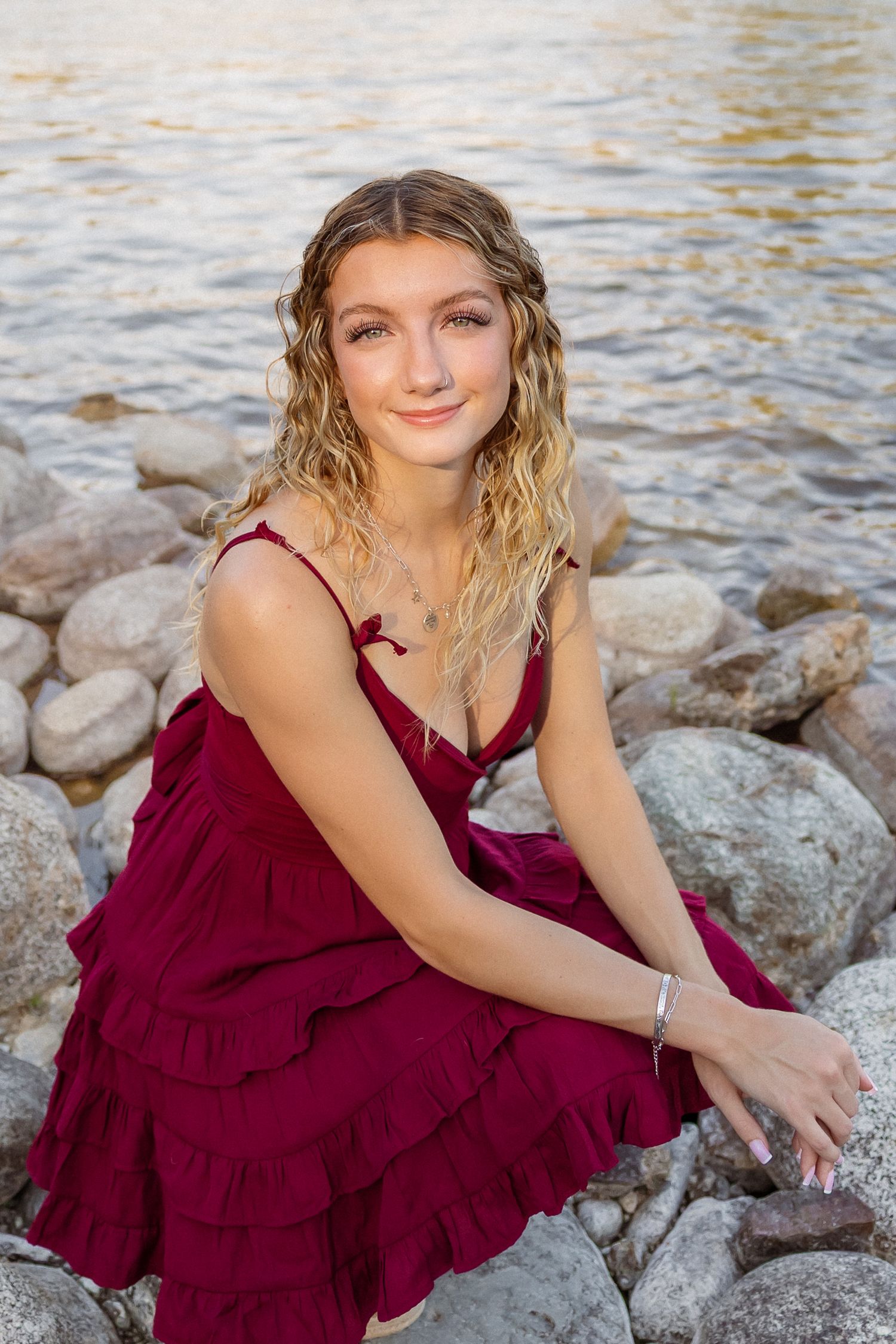 A person in a burgundy ruffled dress poses gracefully on rocky waterfront terrain during a photoshoot.
