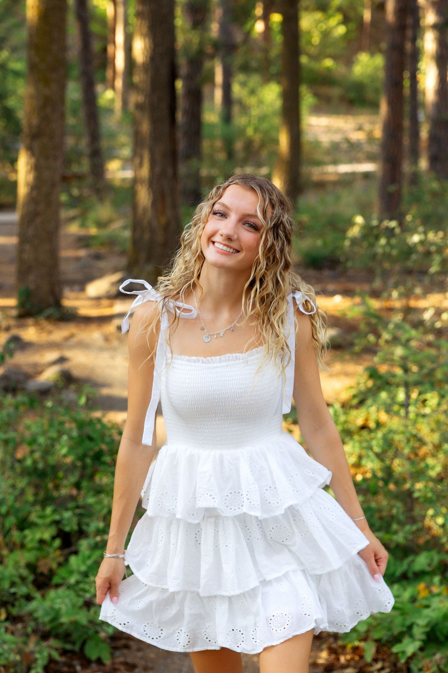 White eyelet cotton sundress with tiered ruffles and shoulder ties photographed in a sunlit forest setting.
