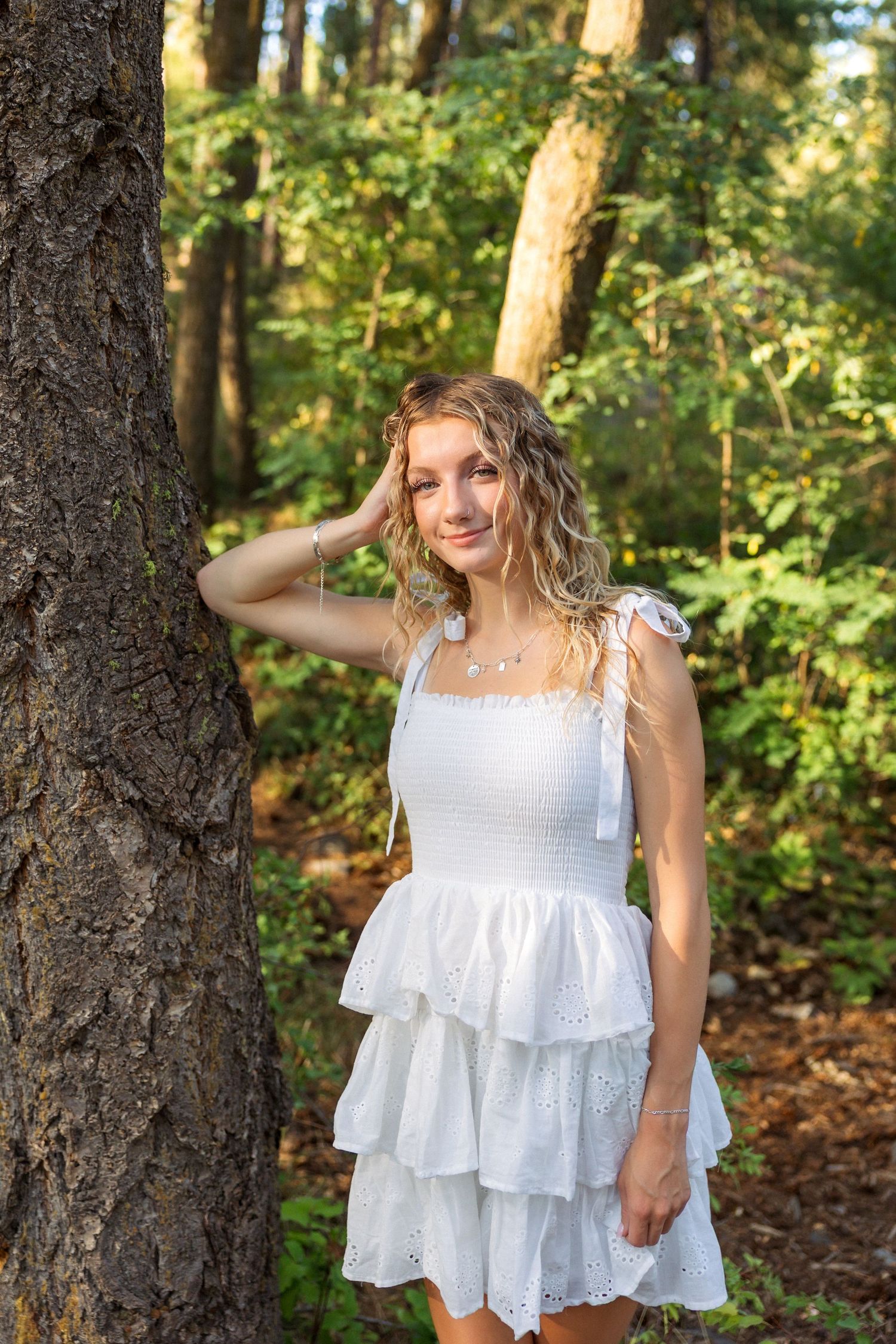 A person in a white tiered summer dress poses next to a tree trunk in a sunlit forest setting.