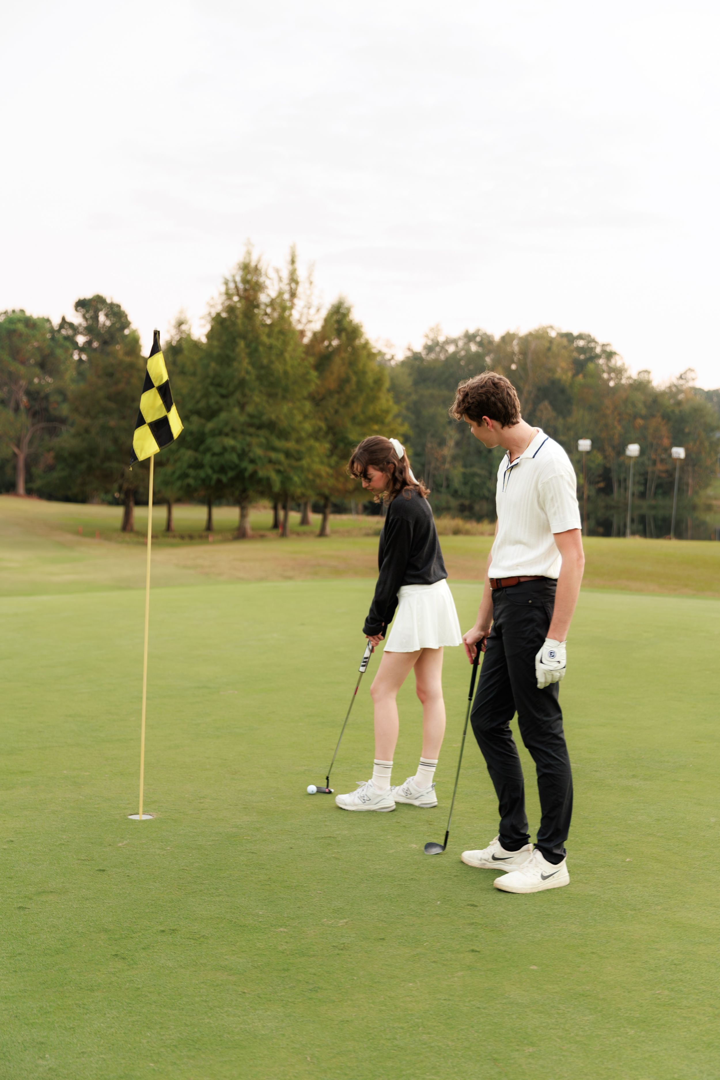 Two golfers practicing putting on a green golf course with a checkered flag pin during late afternoon.