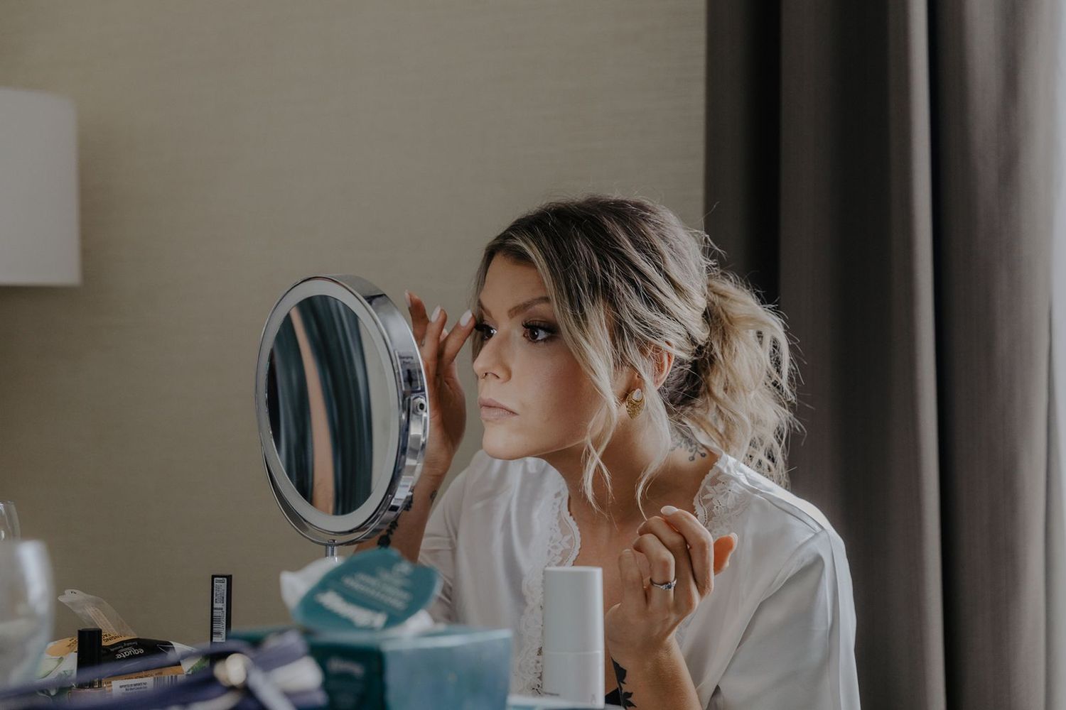 Beautiful bride in white robe checking her makeup in a round mirror at a vanity table before wedding.