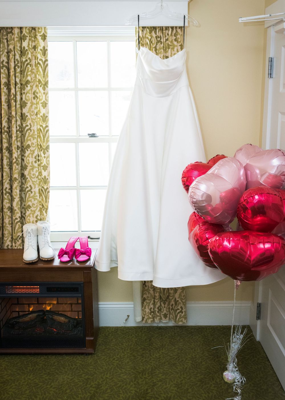 A white dress floats elegantly against a wall with floral curtains and red heart-shaped balloons in a sunlit room.