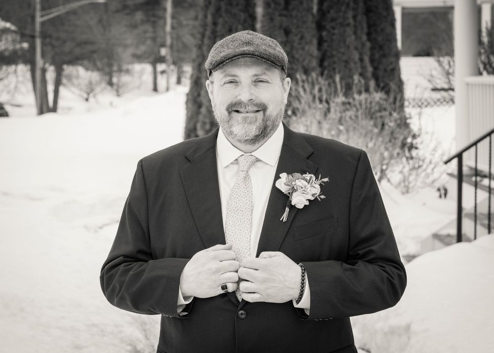 A winter wedding scene showing a person in a black suit holding a beverage in the snow.