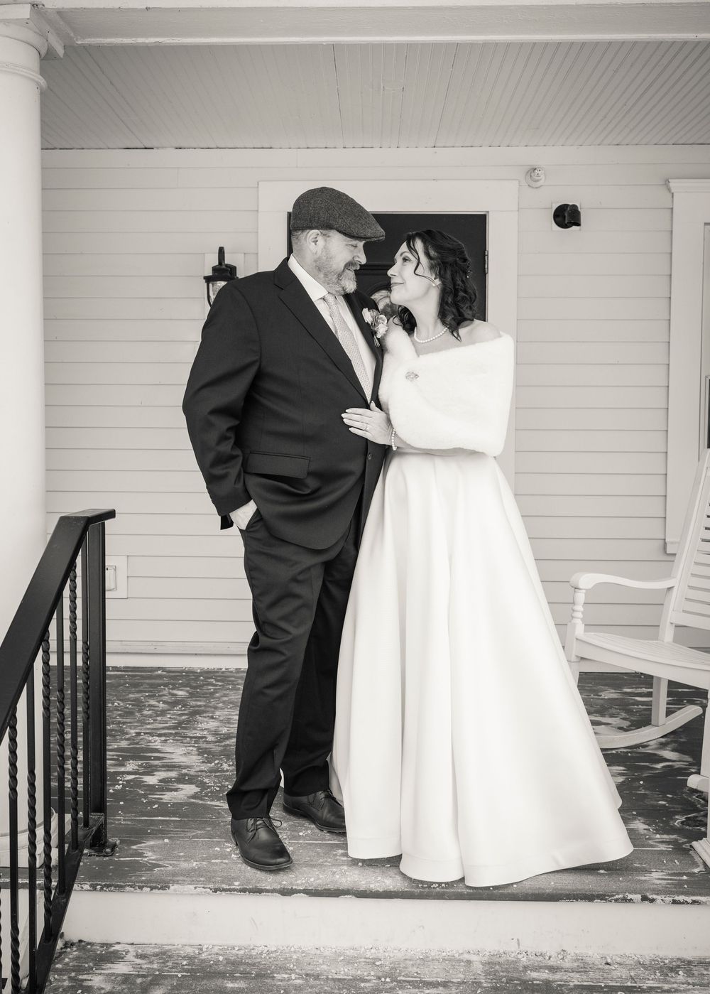 A black and white wedding photo showing a couple sharing a tender moment on a porch with white siding and railings.