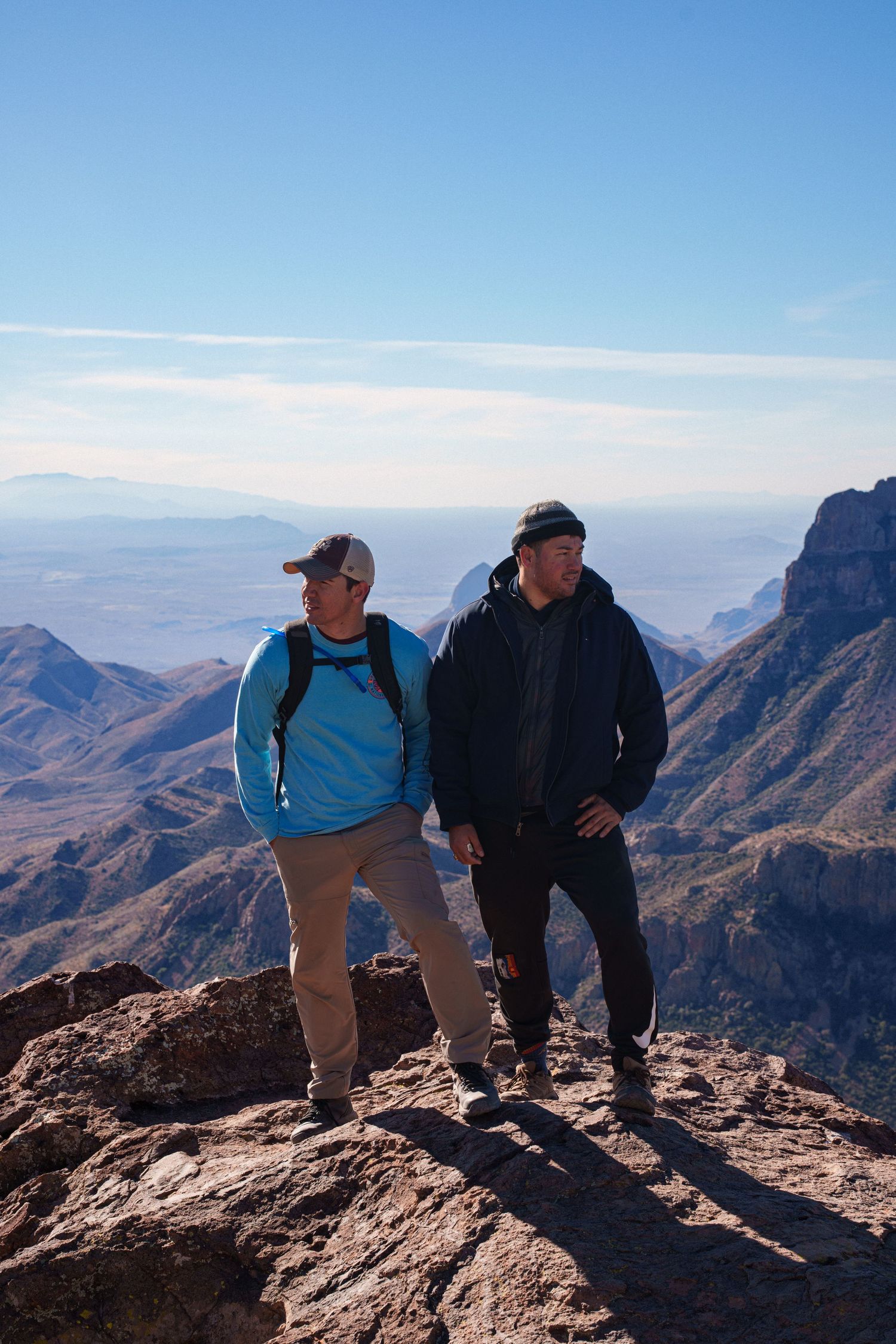 Two hikers stand on a rocky mountain peak overlooking a vast desert landscape on a clear sunny day.