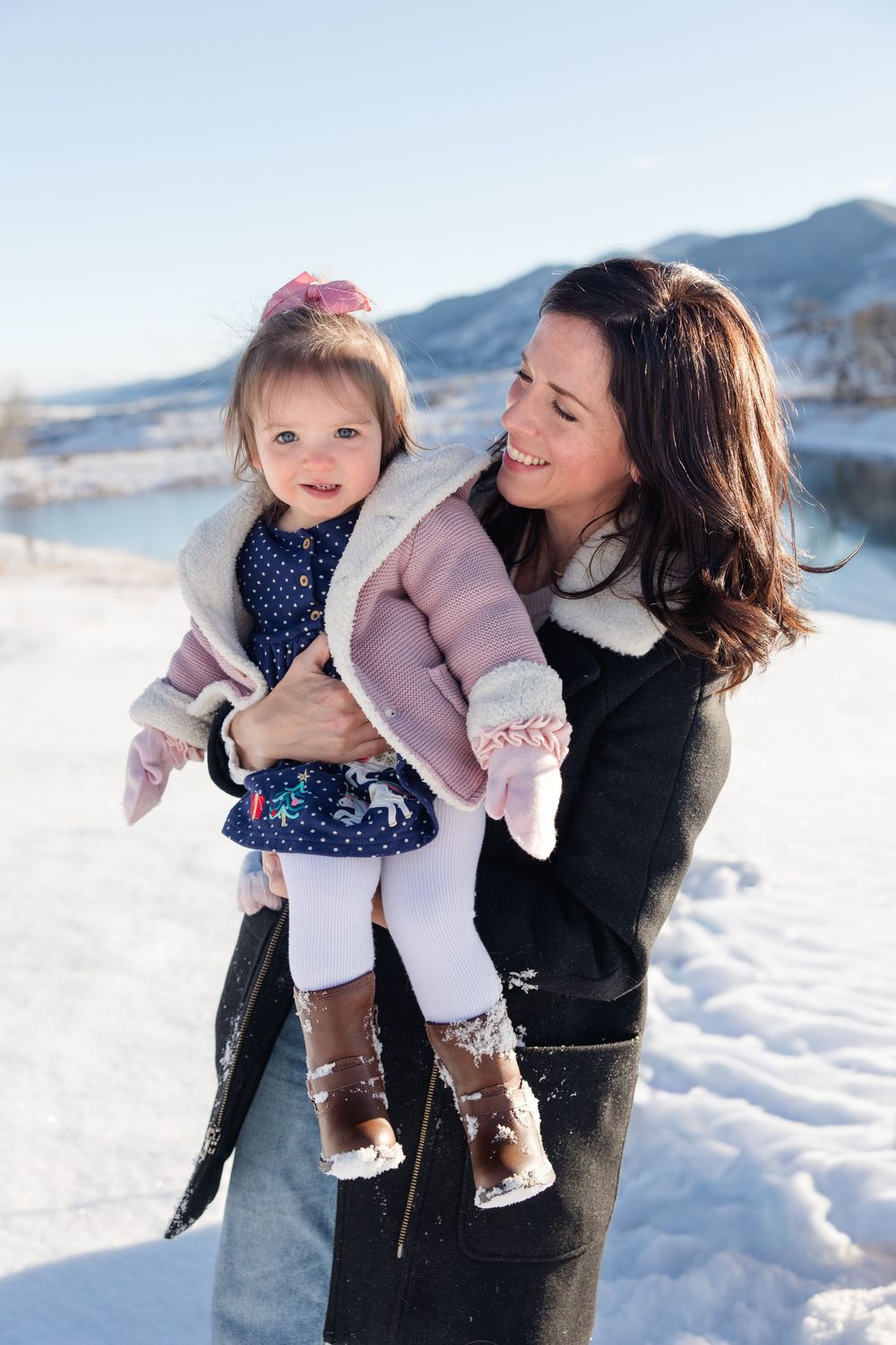 A heartwarming winter photoshoot shows playful moments in the snow with a small child in a pink coat and polka dot dress.