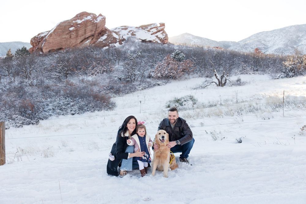 A couple and their golden retriever enjoy a playful moment in the snow with mountains visible in the distance.