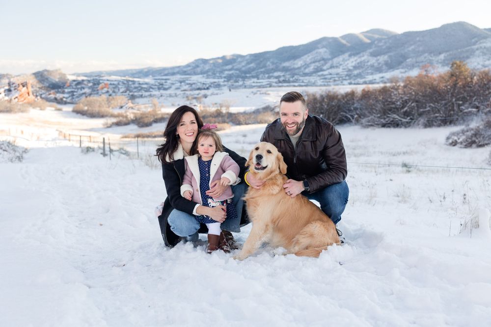 A family and their golden retriever dog pose together in a snowy winter landscape with mountains in the background.