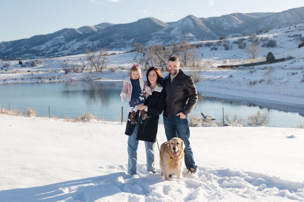 A couple and their golden retriever walk through a snowy winter landscape with mountains in the background.