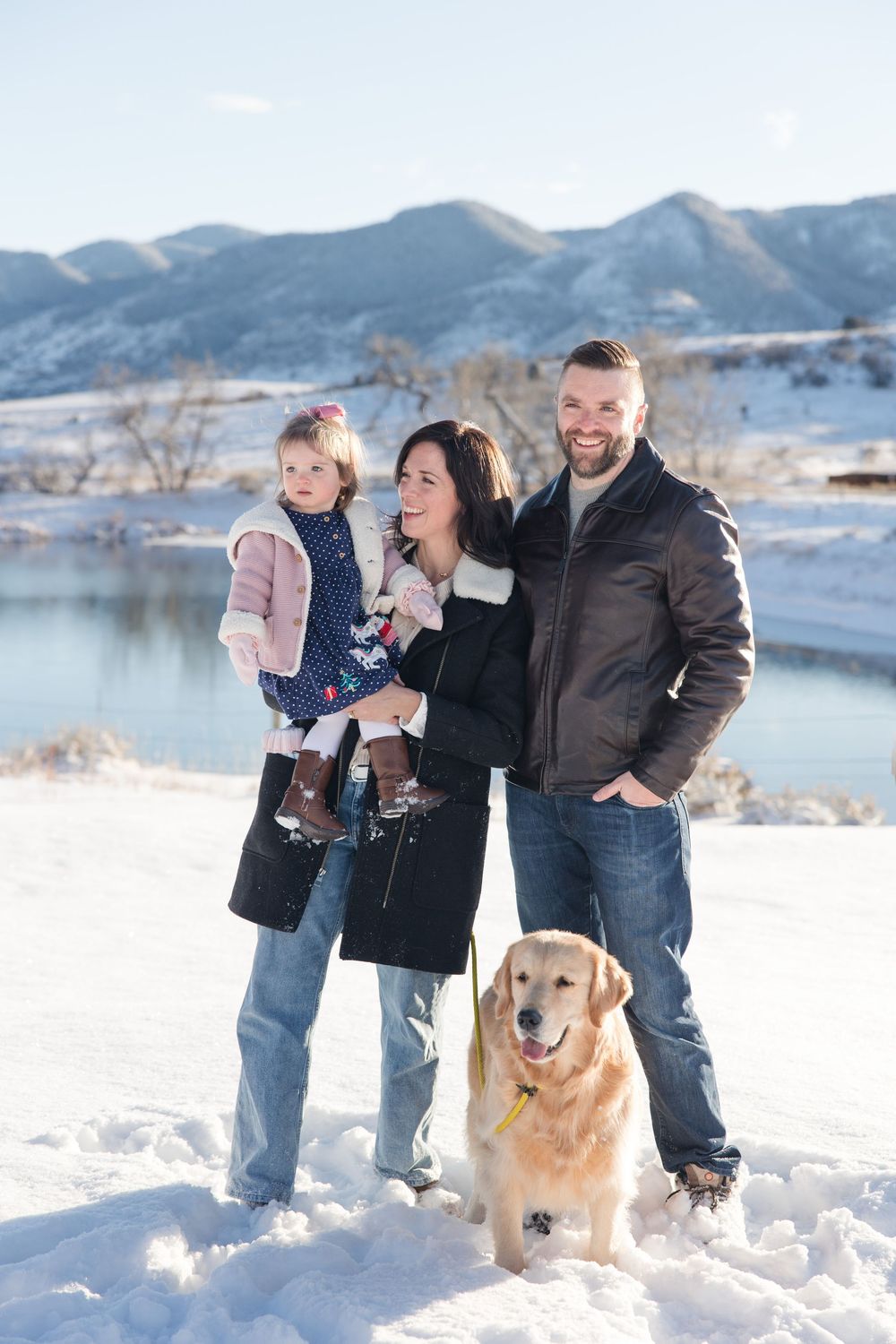 Family and golden retriever playing together in snowy mountain landscape with lake in background.