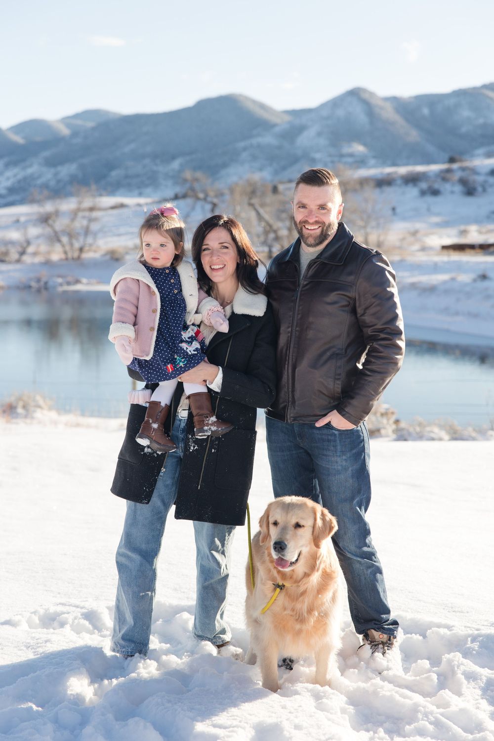 A family and their Golden Retriever pose together in a snowy mountain landscape beside a lake.