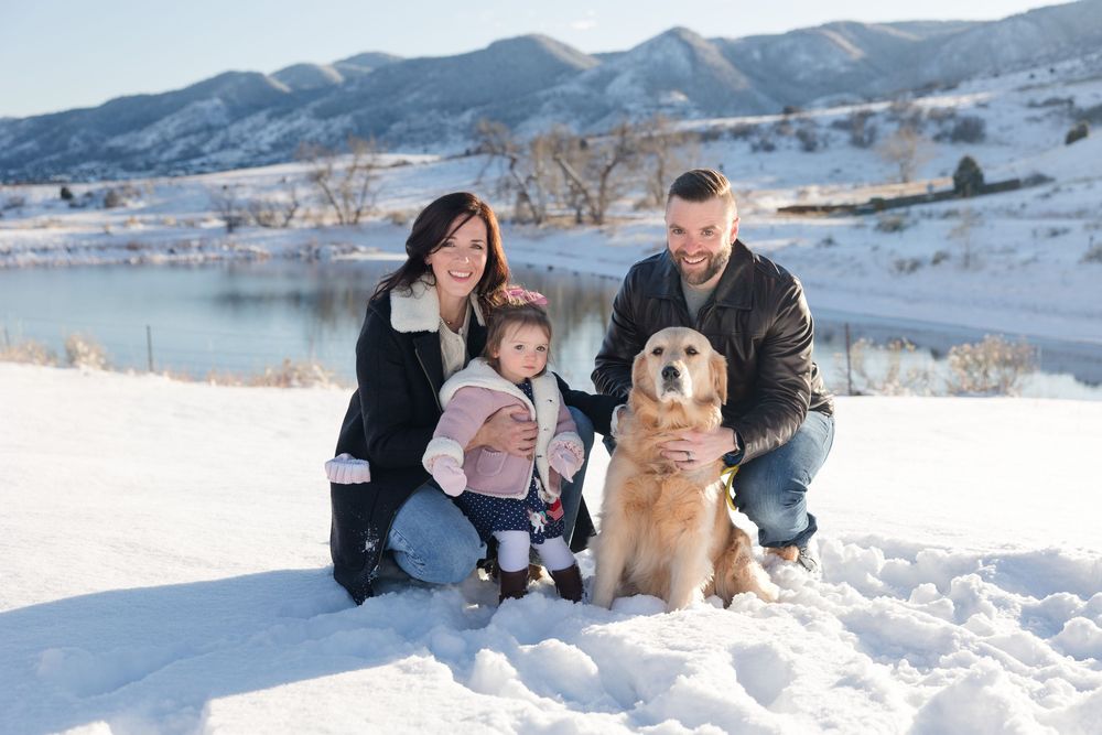 A family kneels in the snow with their smiling Golden Retriever against a mountain backdrop.