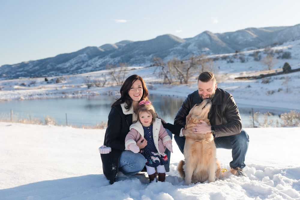 Family and dog playing together in snowy winter landscape with mountains in background.