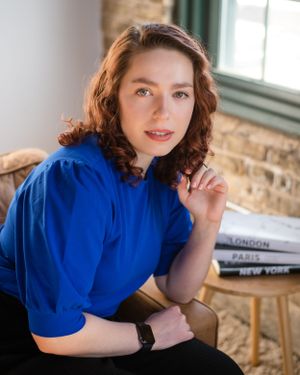 Professional portrait of person in royal blue blouse seated by window with books.