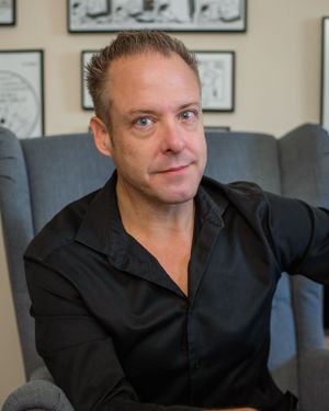 Professional portrait of person in black shirt seated against wall with framed artwork.