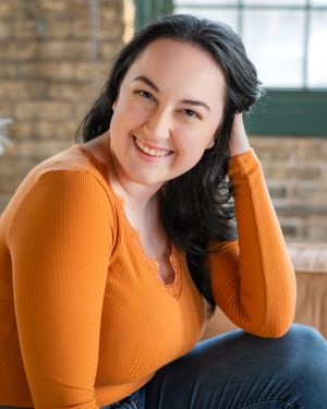 Portrait of someone in an orange sweater posing casually against a brick wall backdrop.
