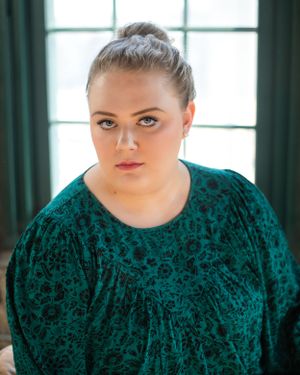 Portrait of person in emerald green velvet dress against window with dark curtains.