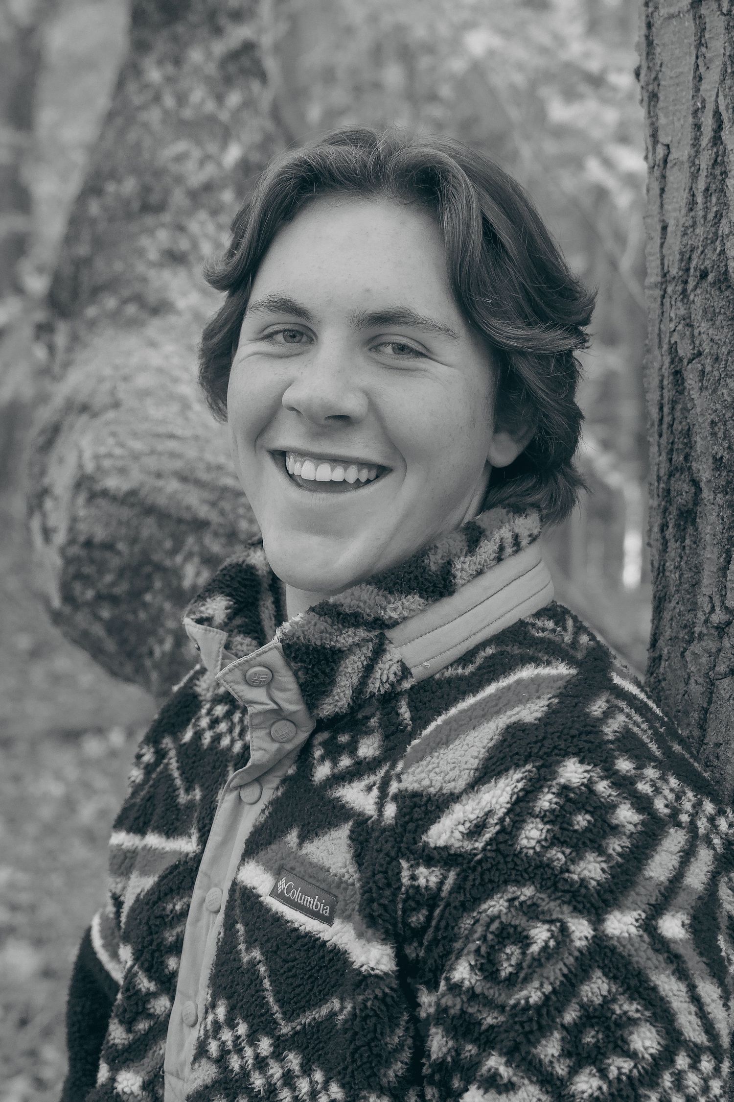 A person wearing a patterned knit sweater smiles while standing against a tree in black and white.