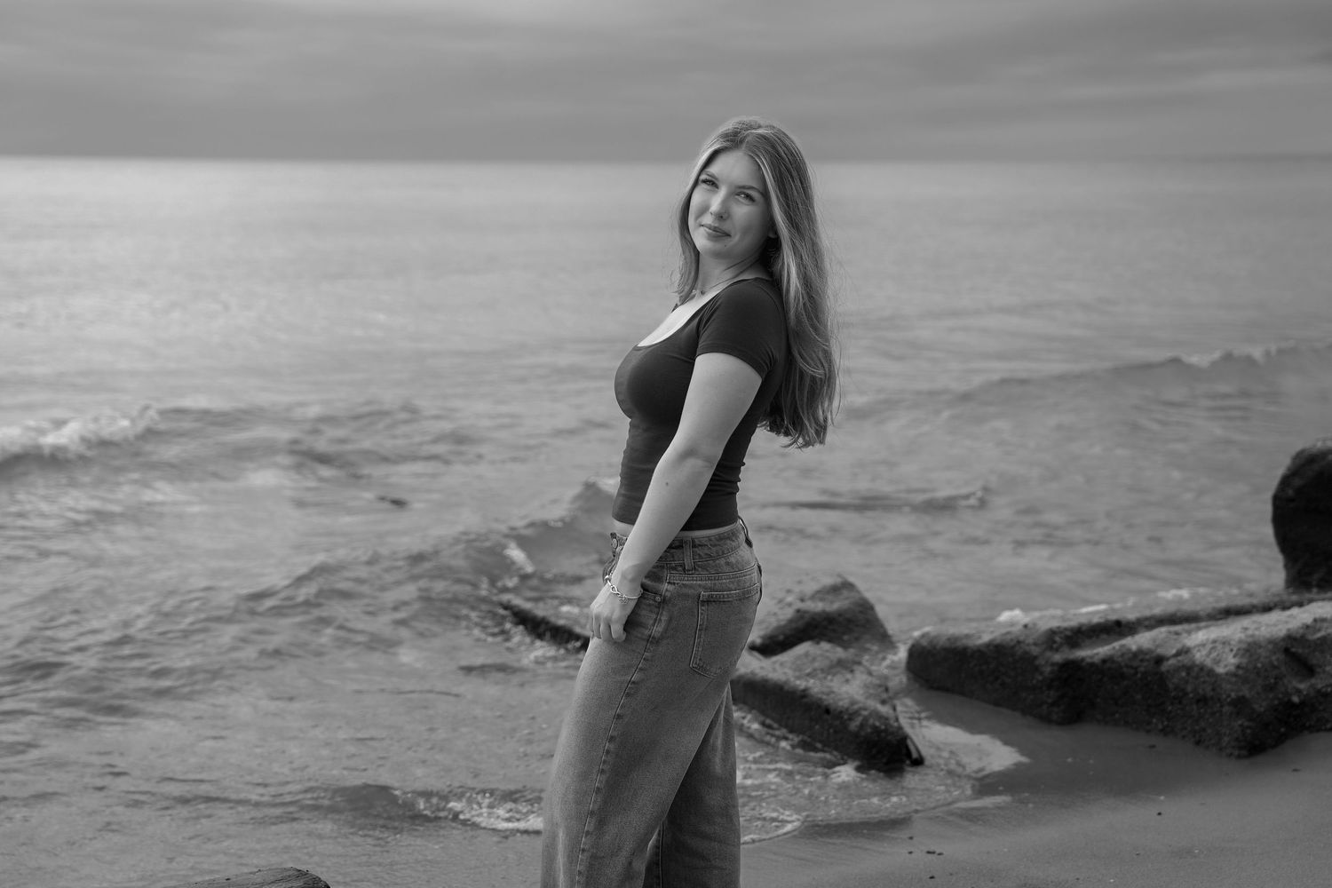 A person in a black swimsuit poses gracefully on rocky coastal cliffs at sunset with ocean waves in the background.