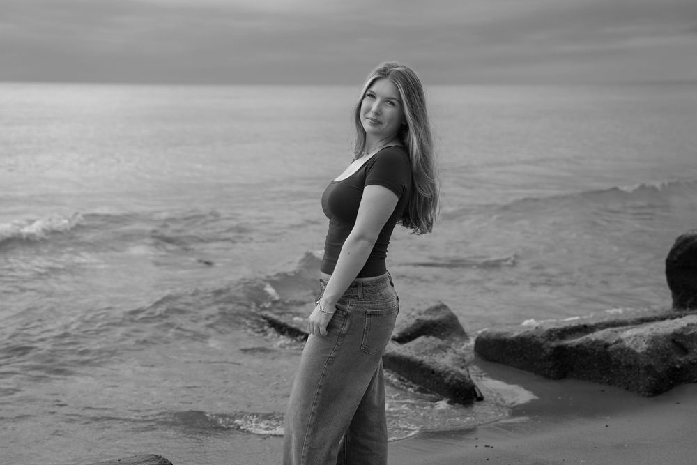 A person in a black swimsuit poses gracefully on rocky coastal cliffs at sunset with ocean waves in the background.